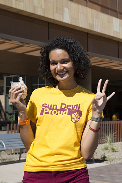 A young lady in a gold shirt reading "Sun Devil Proud" holds up an ice cream sandwich and ASU's hand symbol, the pitchfork. 