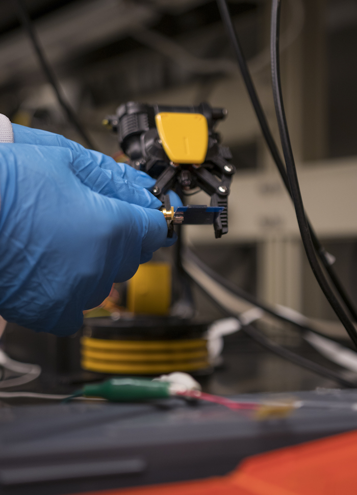 Photo of a pair of hands in blue gloves working with equipment. Caption: The brain implant prototype is held next to an external antenna that emits electromagnetic waves to the internal device. The electromagnetic waves reflect back with the brain signal patterns for external analysis. Photographer: Marco-Alexis Chaira/ASU