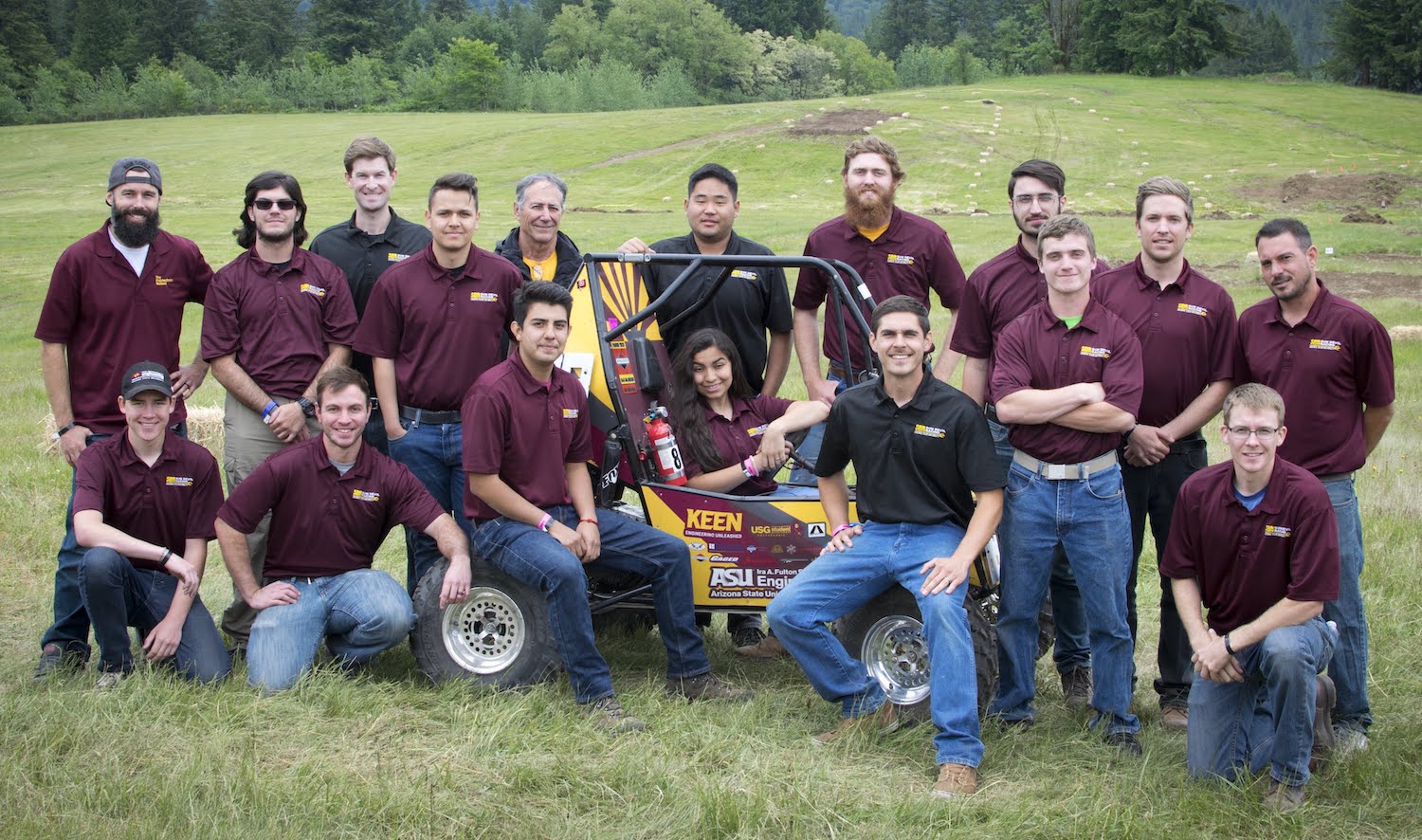 Photo of a large group of people with a race car in a field.