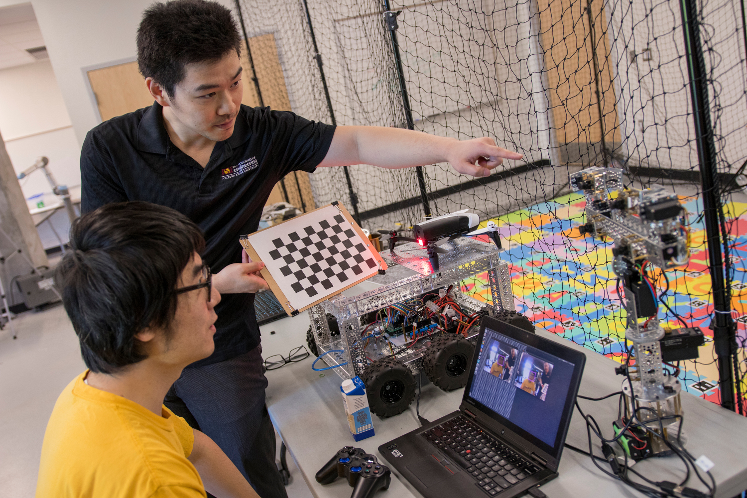 Professor standing next to seated student pointing at a structure that features cameras